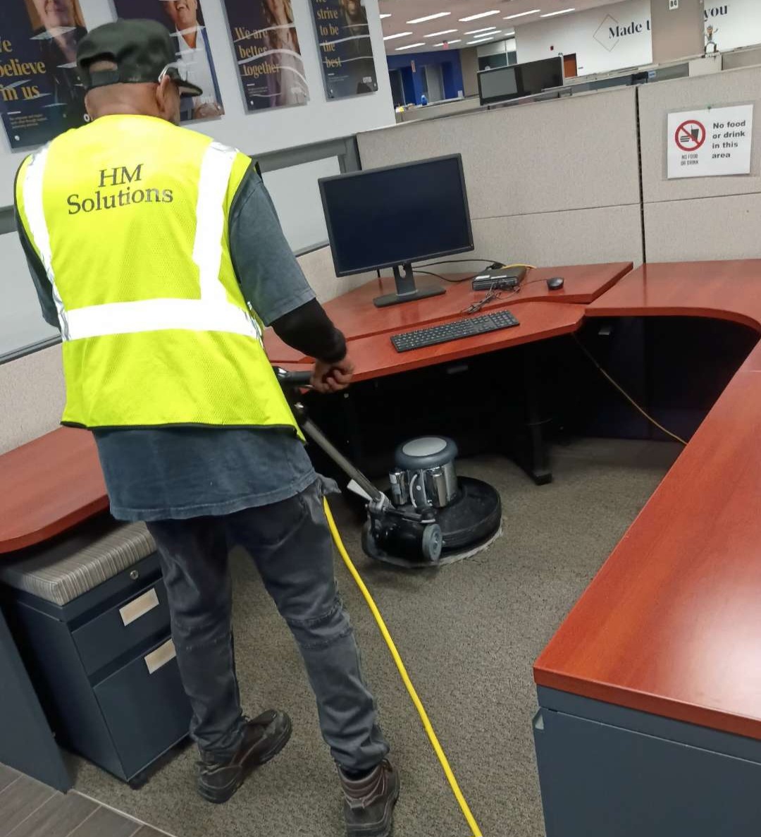 An HM Solutions crew member in a safety vest running a floor buffer through an office cubicle area