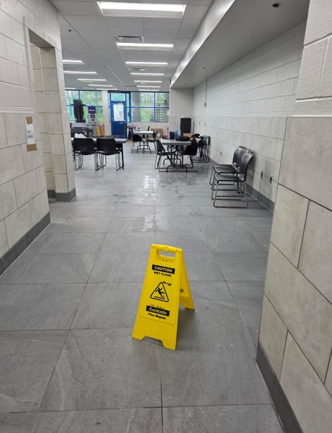 A plant canteen and corridor at an HM Solutions account, freshly mopped with a bilingual caution wet floor sign in place