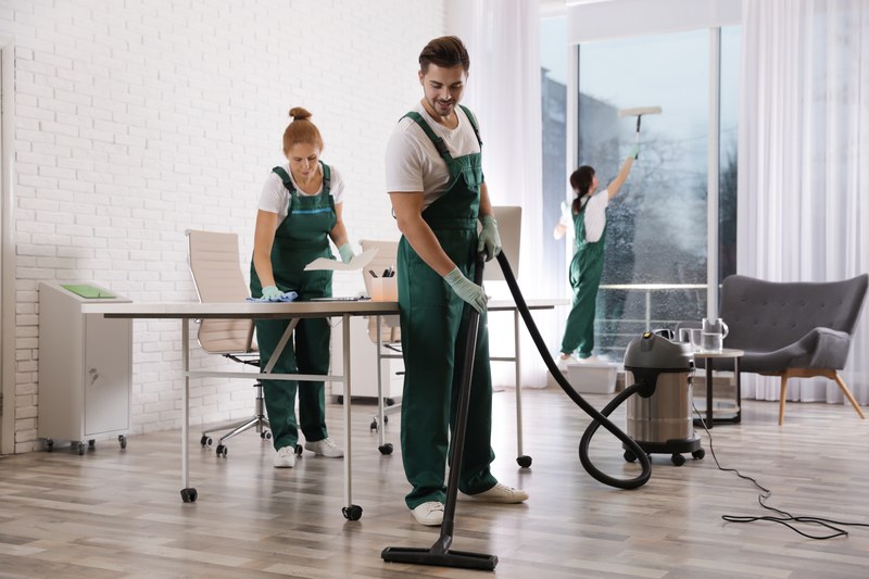 A three person cleaning crew working an office together, vacuuming, wiping and cleaning glass