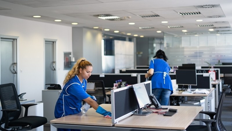 Cleaning staff working through an open plan office, wiping desks and workstations